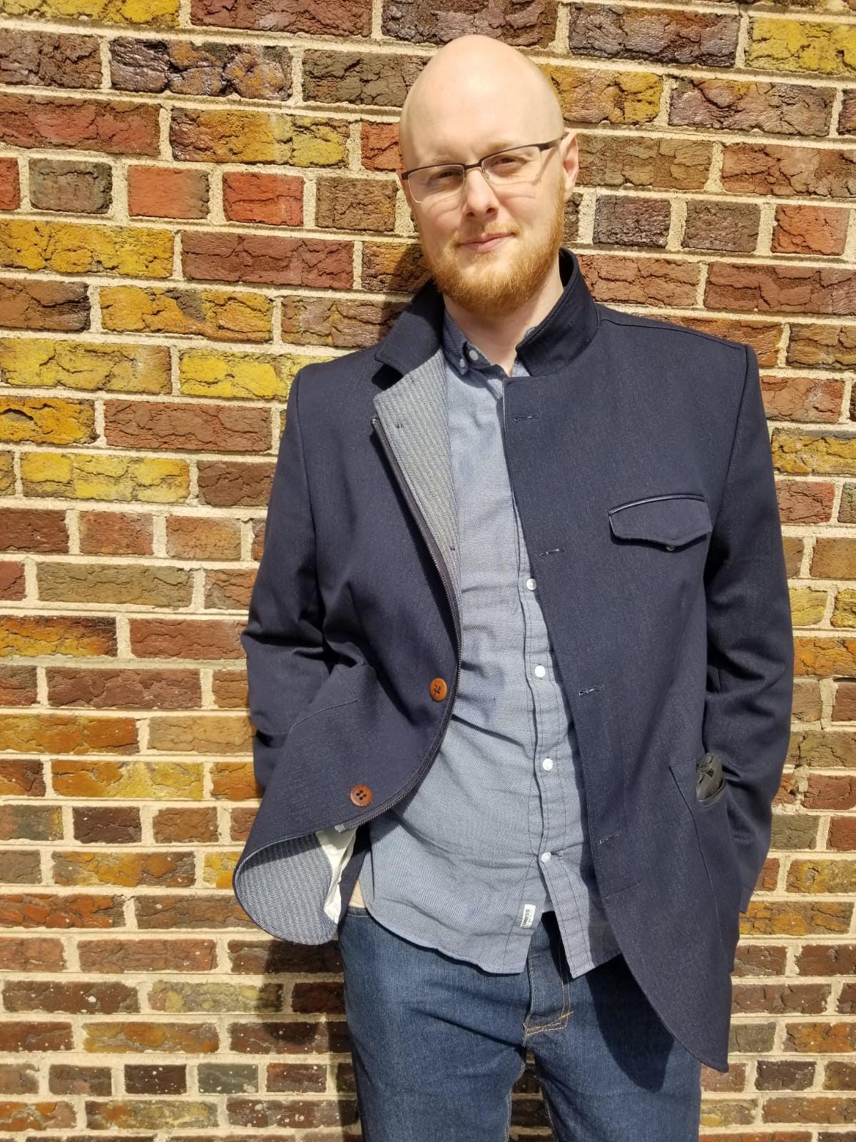 Rex Barkdoll in a dark blue jacket, grey button down shirt and jeans against a brick wall